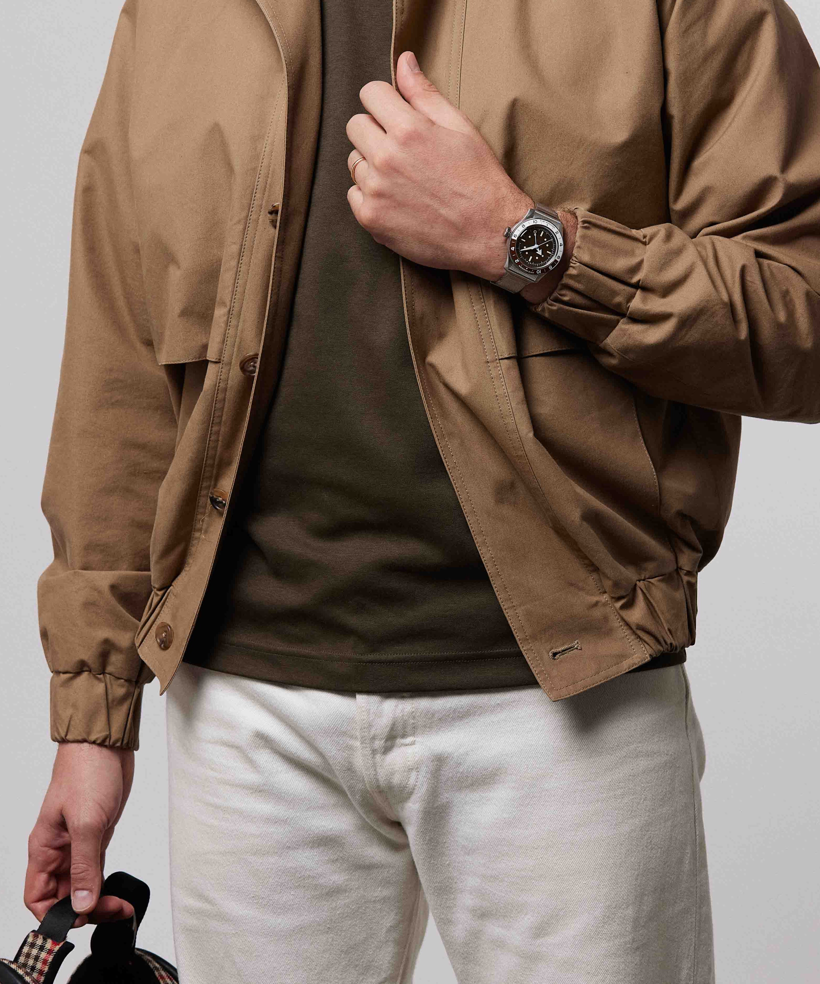 Serica Watches - Homme debout tenant sa veste beige portant la montre SERICA 8315-2 GMT marron en acier étanche 200m.