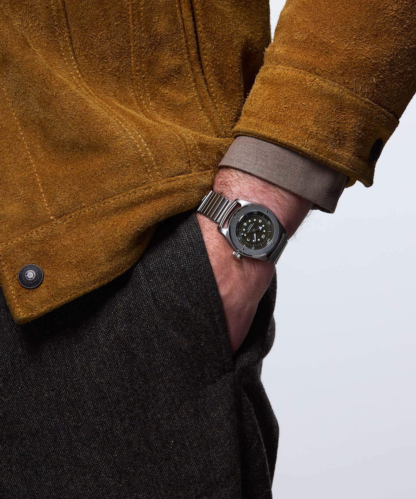 Serica Watches - Chronomètre étanche SERICA 7505-2 Vert Olive sur bracelet acier Bonklip, porté au poignet d'un homme habillé d'un blouson en cuir suédé daim et d'un pantalon en laine texturée marron, la main glissée dans la poche.